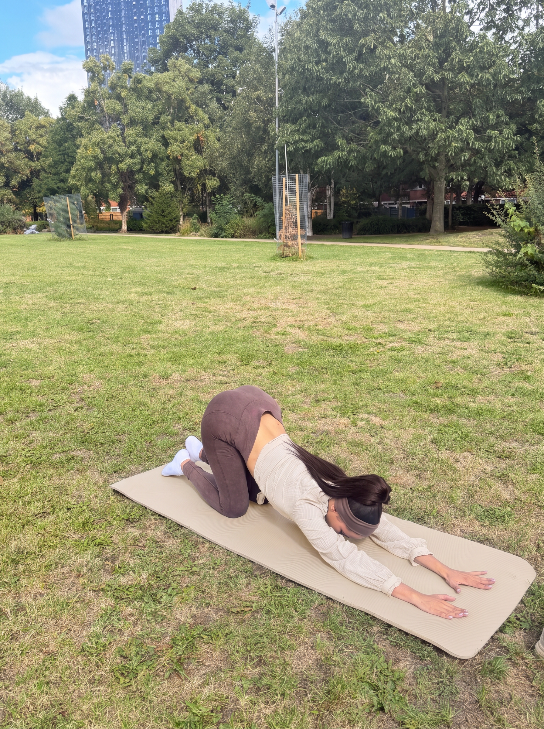 Ana in a beige shirt stretching in child's pose on a beige yoga mat in the park